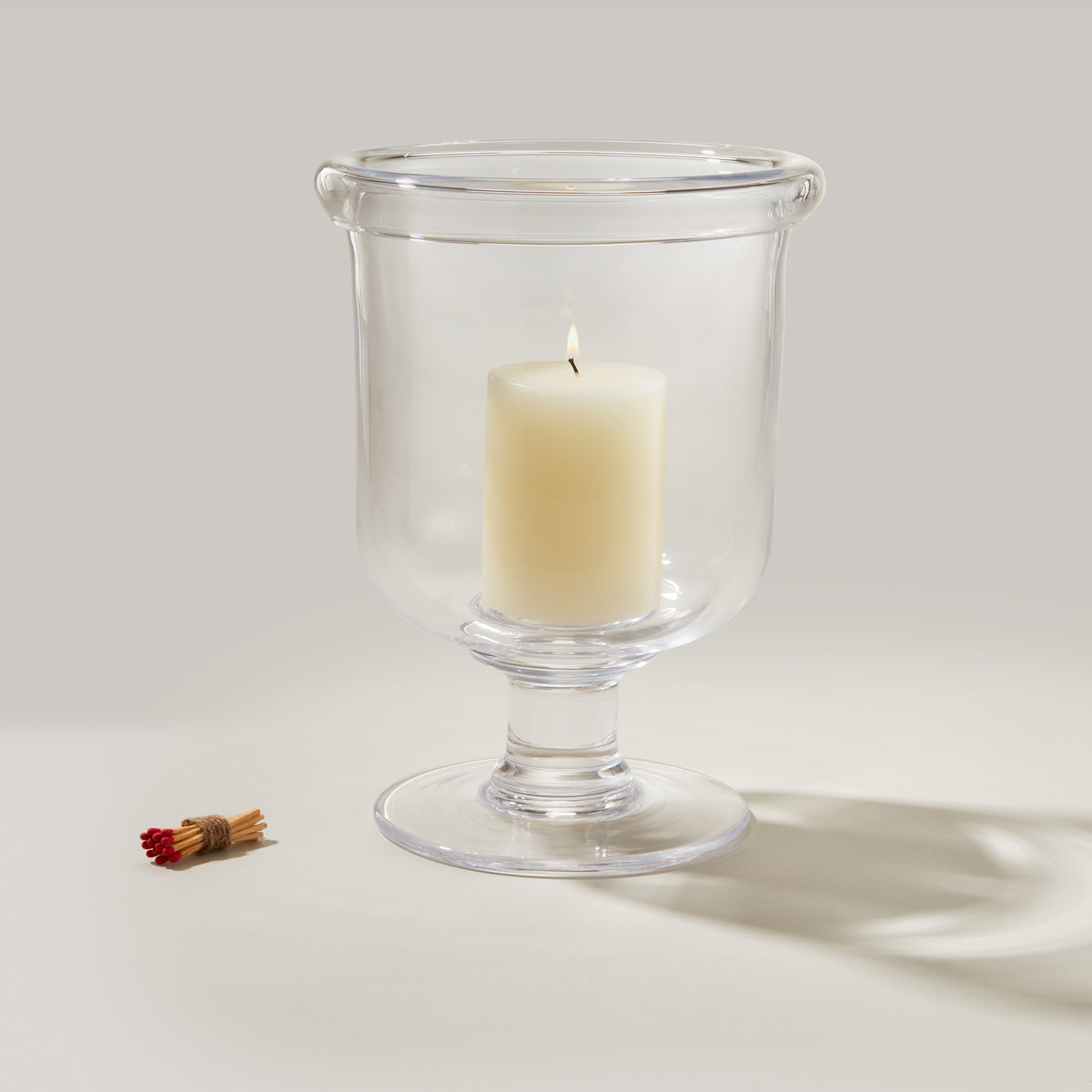 Clear glass hurricane lamp with cream pillar candle inside and red matchstick bundle beside it.