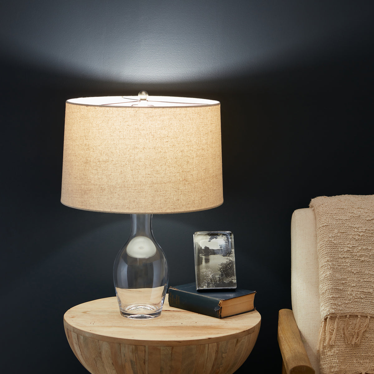 Table lamp with clear glass base, white interior, and tan linen drum shade, displayed on wooden round table.