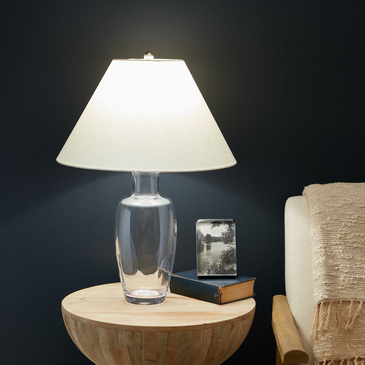 Tall lamp with clear glass vessel base and cream trapezoid linen shade, illuminated on wooden round table.