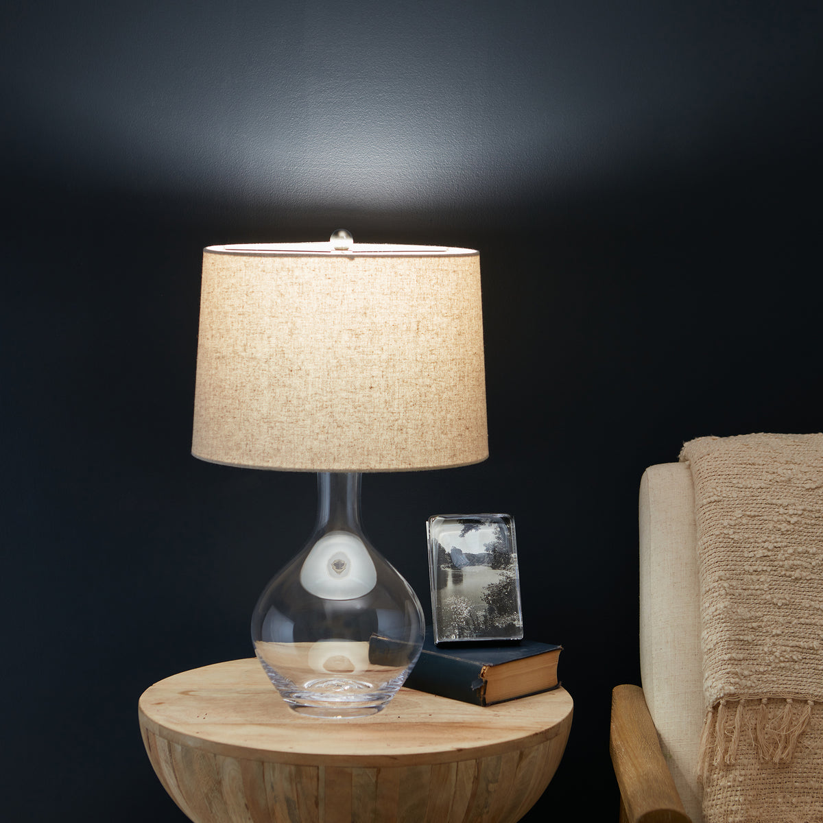 Glass table lamp with beige linen shade and round bulbous base on wooden round table.