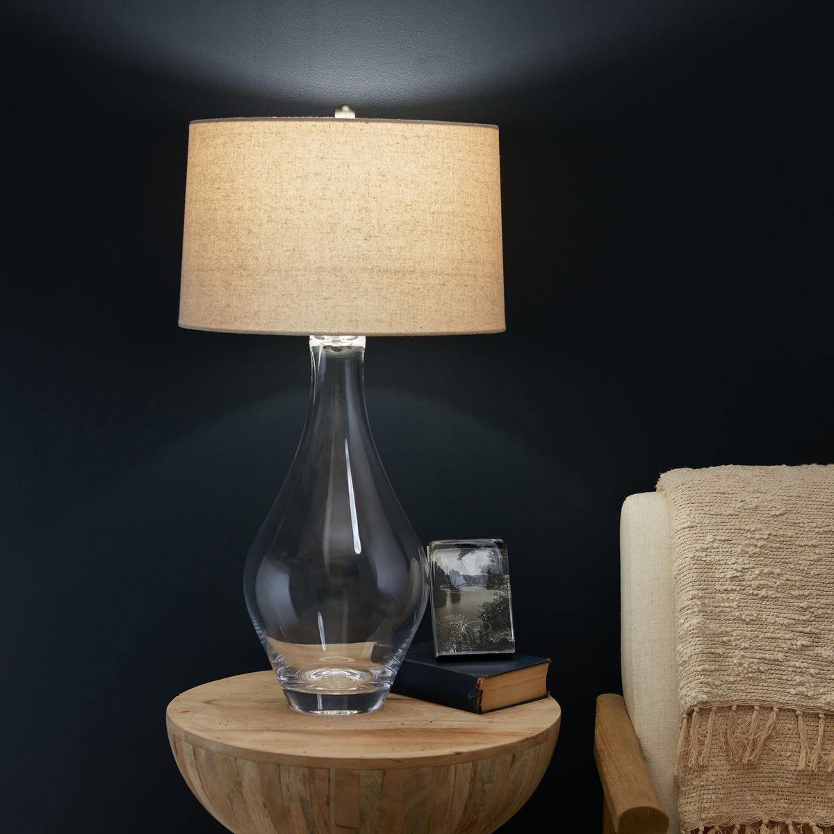 Clear glass teardrop-shaped lamp base with tan linen drum shade on wooden side table.