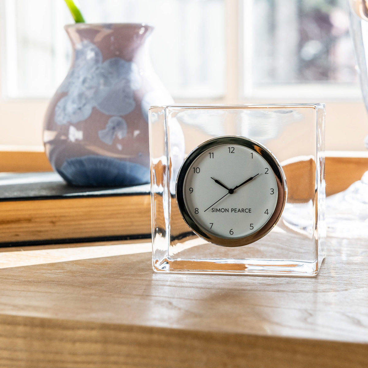 Clear glass cube desk clock with black frame and white dial showing Simon Pearce branding.