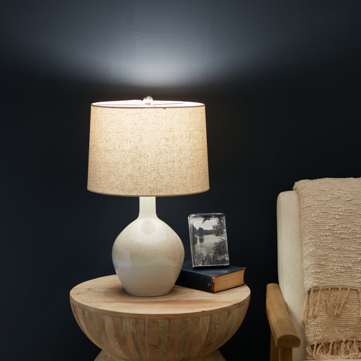 Cream ceramic table lamp with textured glaze, round base, and linen shade on wooden side table.