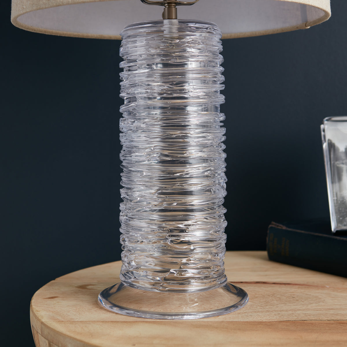 Clear textured glass cylindrical lamp base with linen shade on wooden surface.
