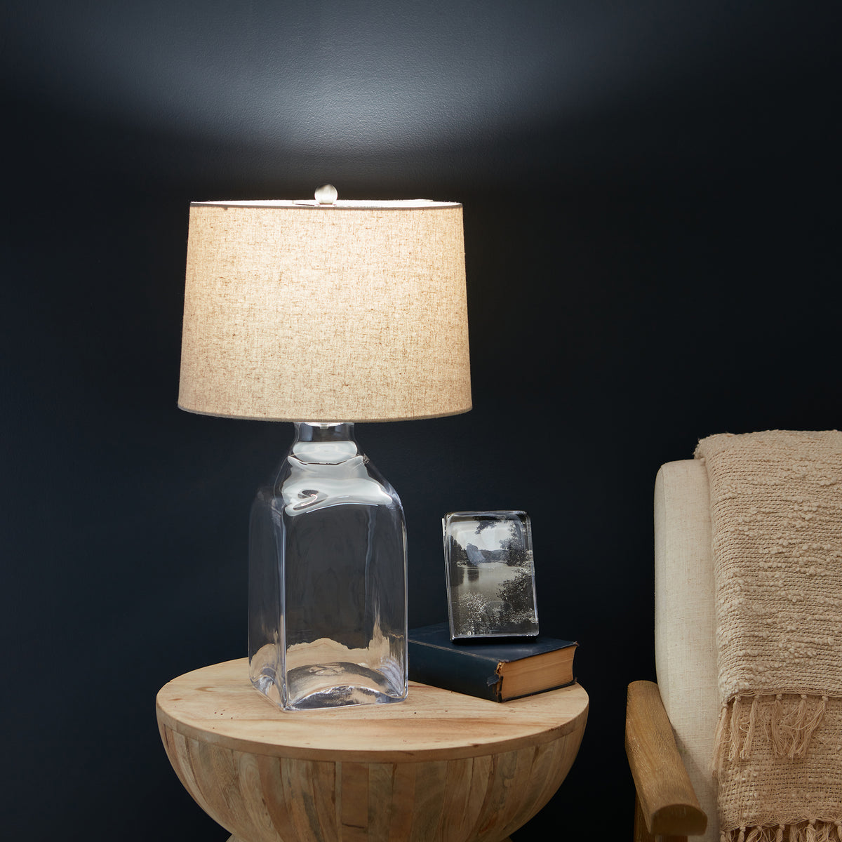 Glass bottle table lamp with beige linen drum shade and white finial, displayed on wooden bowl table next to furniture.