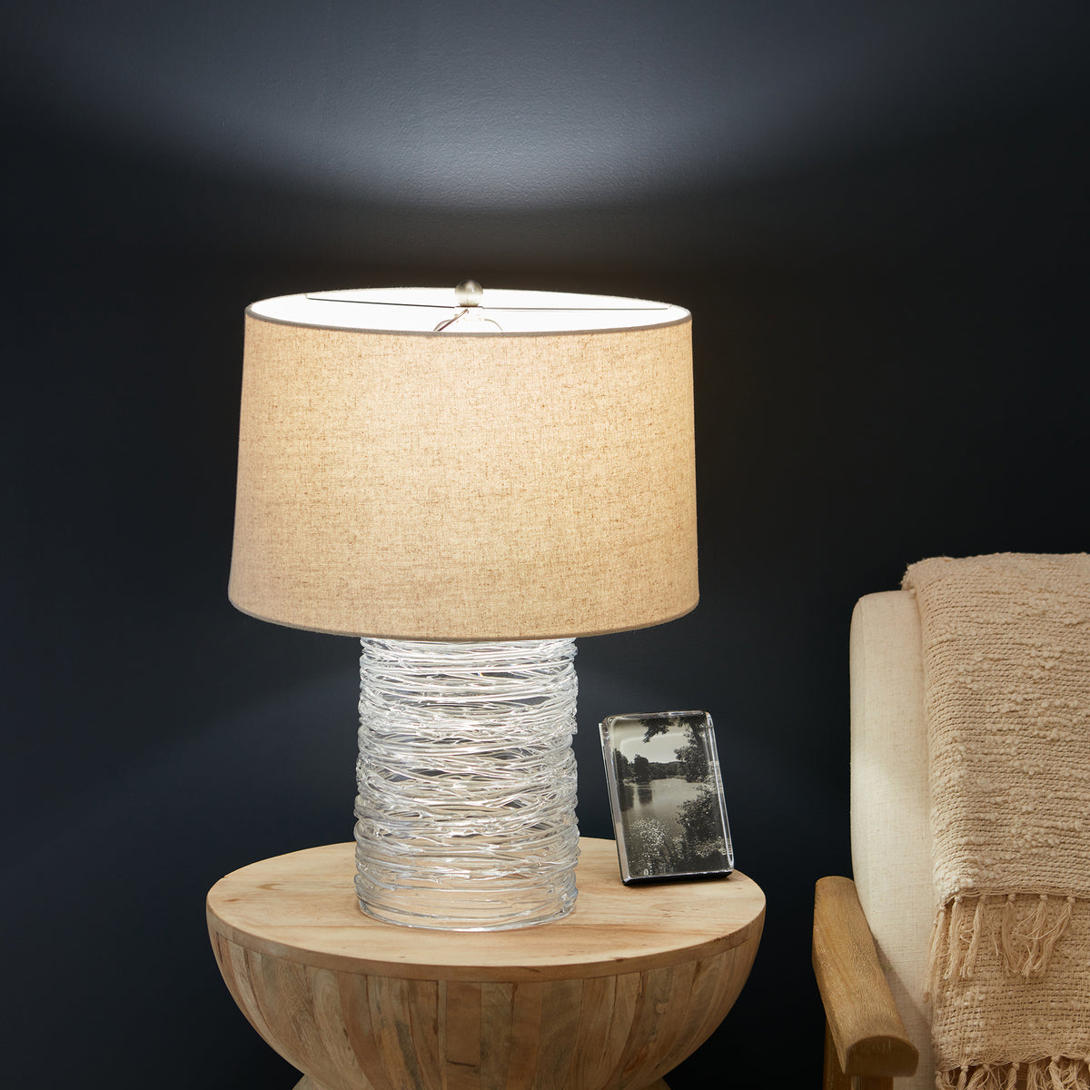 Cylindrical table lamp with textured white ceramic base and tan linen shade on wooden pedestal table.
