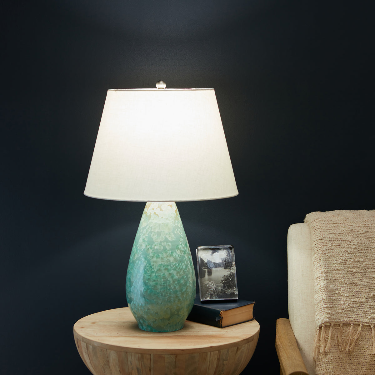 Teardrop-shaped crystalline base lamp in green and cream with white linen shade on wooden table.