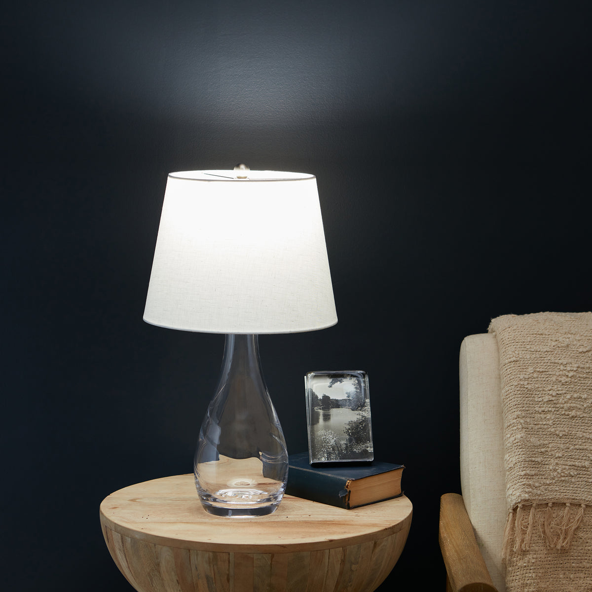 Clear glass teardrop-shaped lamp base with white linen shade on wooden side table.