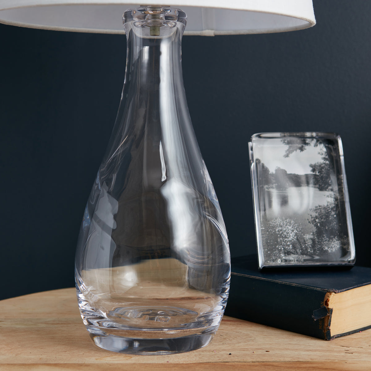 Clear glass bottle-form lamp base with cream linen shade on wooden surface.