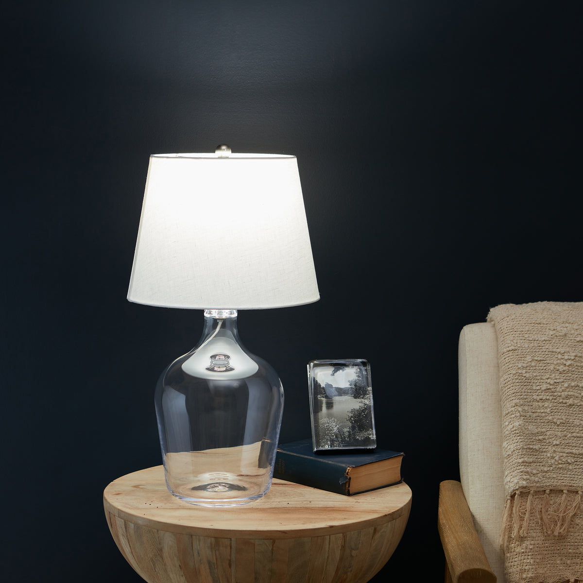 Glass bottle table lamp with white linen shade on wooden round base, next to stacked books and photo frame.