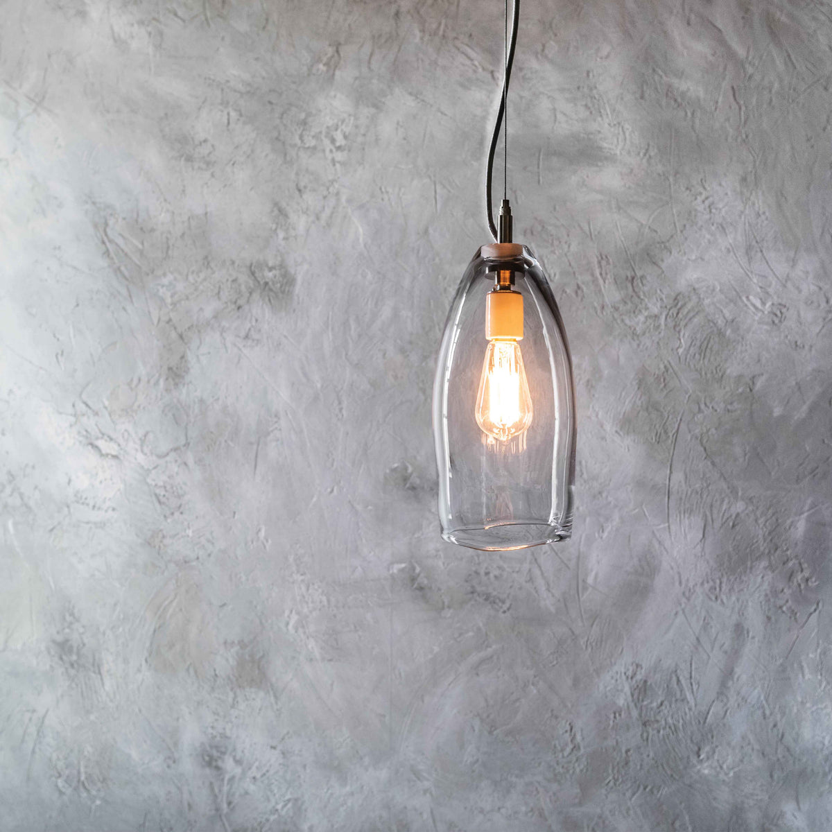 Tall clear glass pendant light with Edison-style bulb and copper fixture hanging from black cord against textured wall.