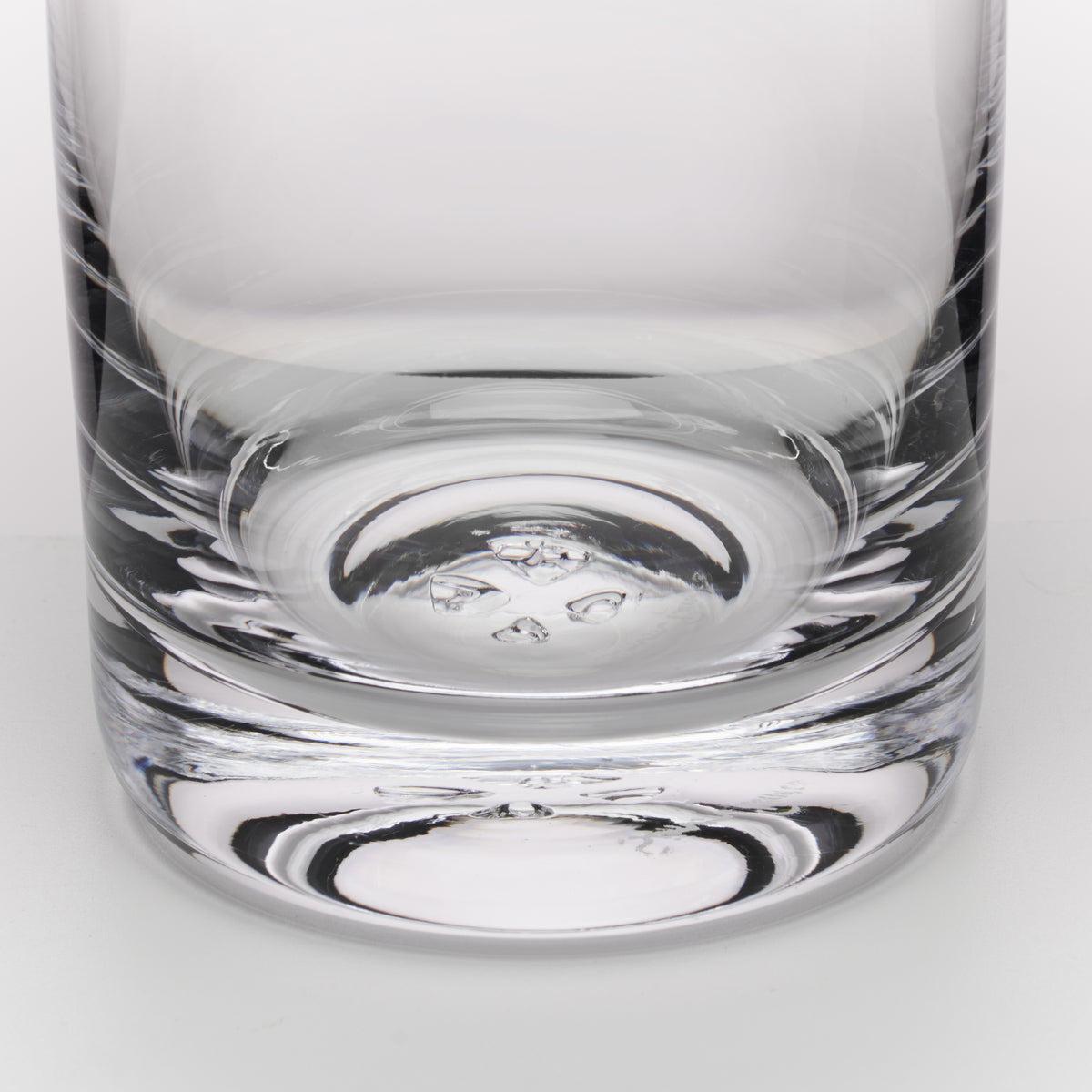 A close-up of a clear glass tumbler with a thick, textured base, showing reflections on a light gray background.