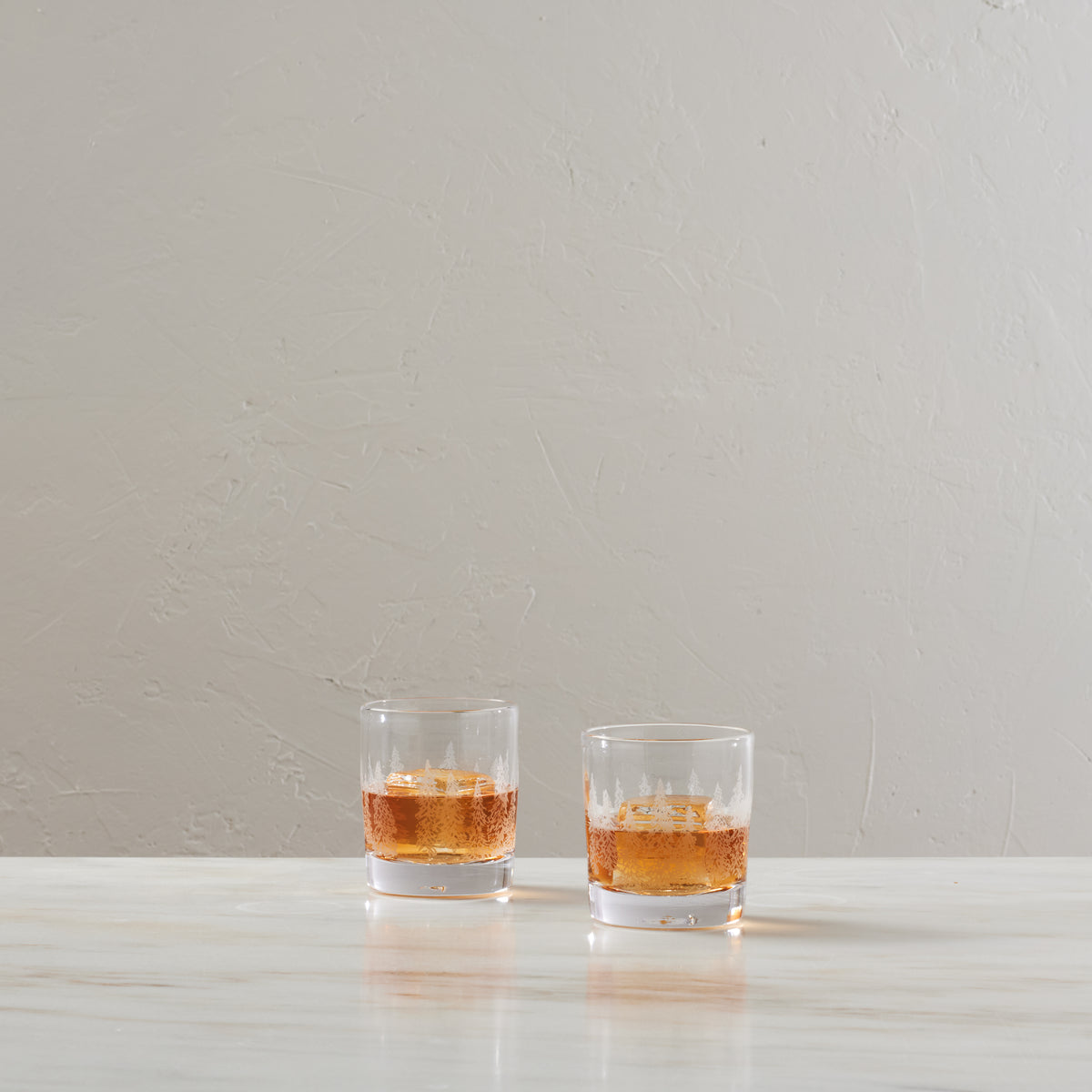 Two clear double old-fashioned glasses with engraved forest design, each holding amber liquid with ice.