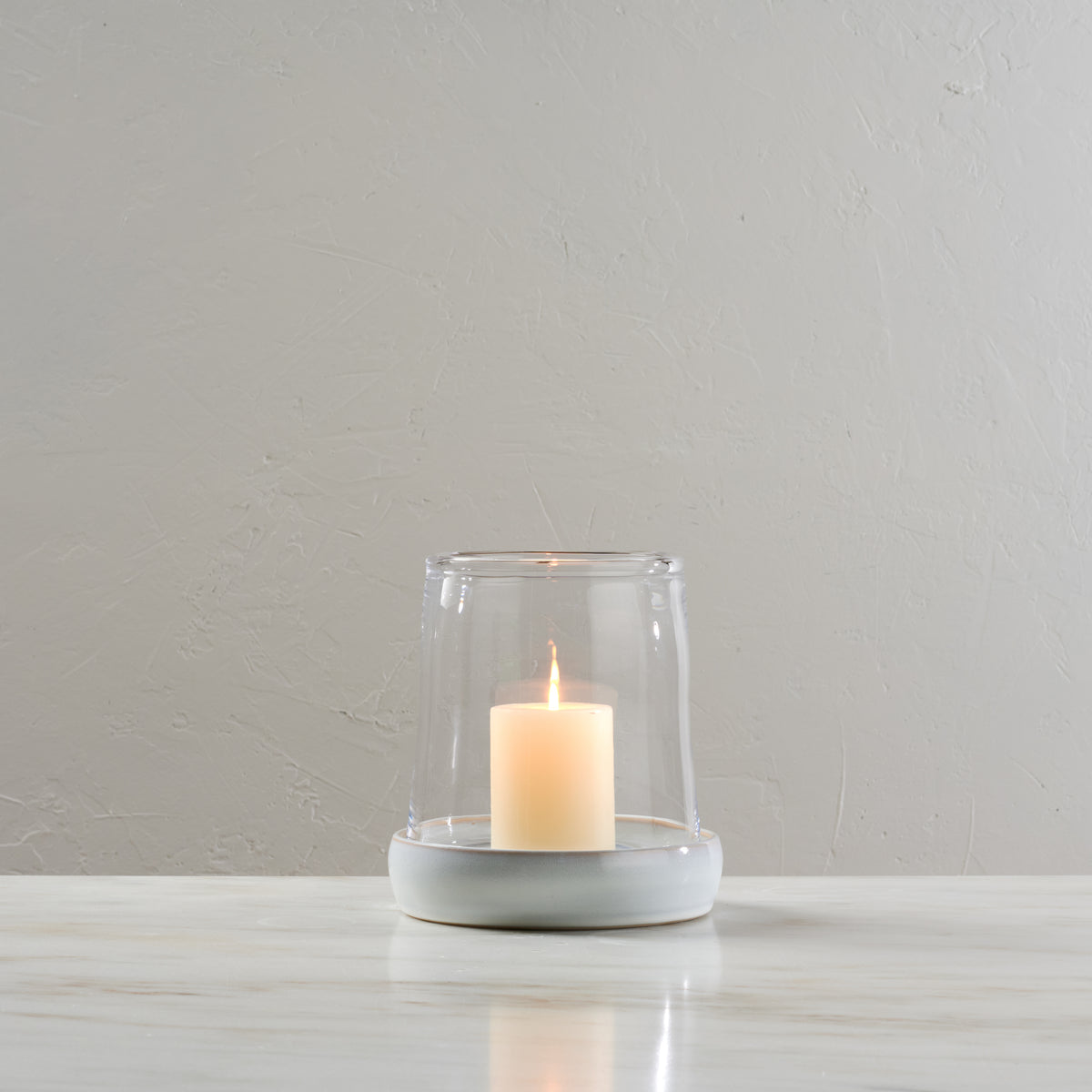 Large hurricane candle holder with glass cylinder and light gray ceramic base holding a lit cream-colored pillar candle.