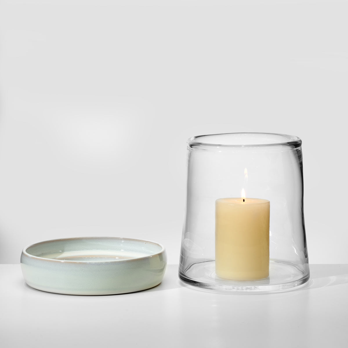 Clear glass hurricane with cream-colored pillar candle inside, paired with a pale green ceramic base dish.