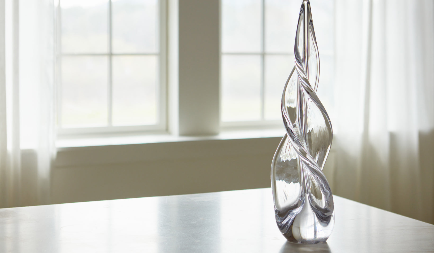 Simon Pearce handblown spiral glass sculpture on a white surface in front of a bright window with sheer curtains