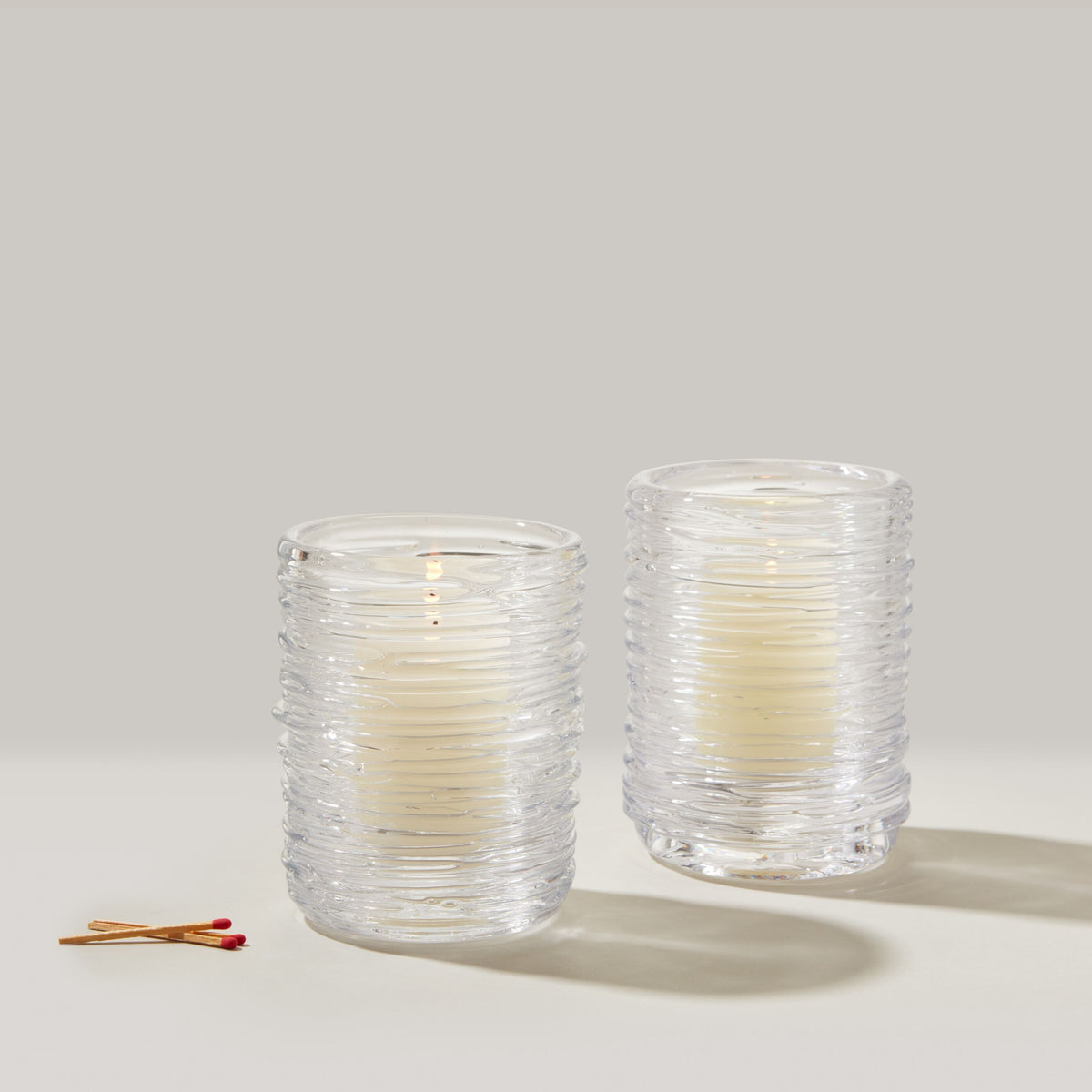 Two clear textured glass hurricane candles with cream-colored wax and matches on neutral background.