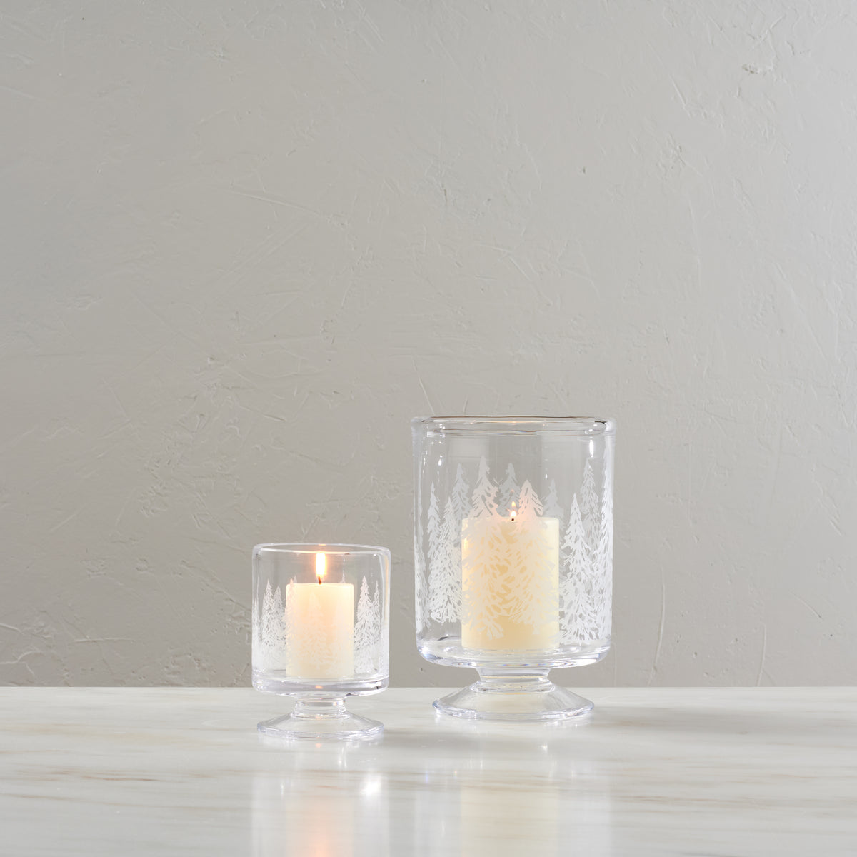 Two clear glass hurricane candle holders with engraved forest tree design, one smaller and one larger, each containing a lit 