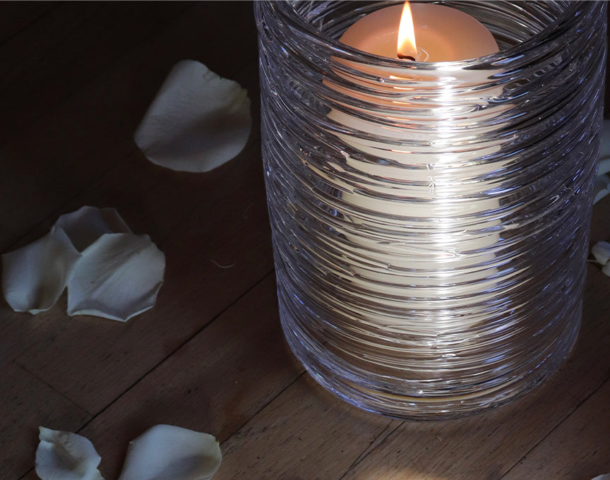 A Simon Pearce ribbed glass hurricane holding a lit pillar candle, surrounded by scattered white rose petals on a dark wood surface