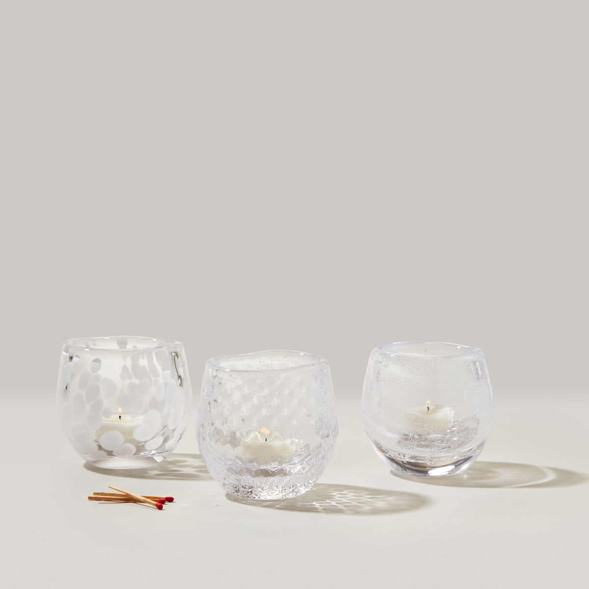 Three clear glass tealight holders with textured surfaces containing white tealight candles, displayed with matches.