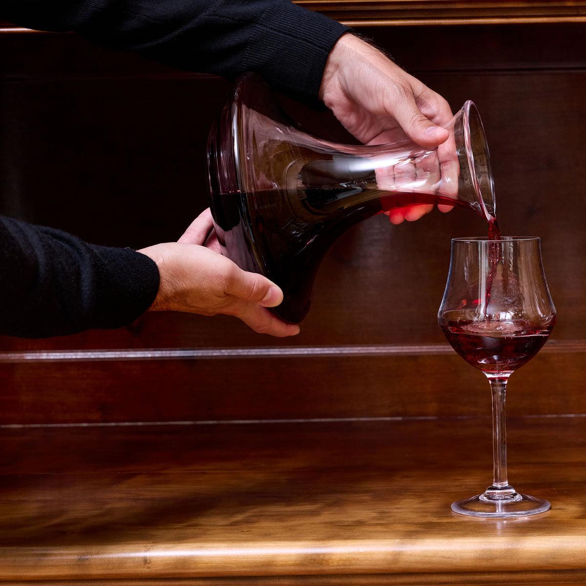 Hand pouring red wine from a clear glass decanter into a wine glass on a wooden surface.