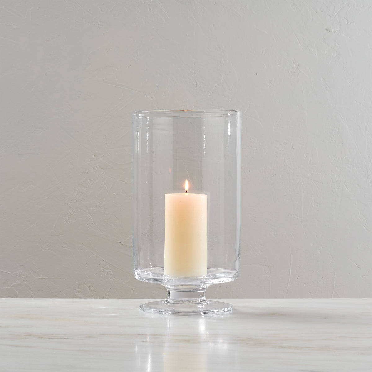 Clear glass hurricane candleholder with pedestal base holding a lit cream-colored pillar candle.