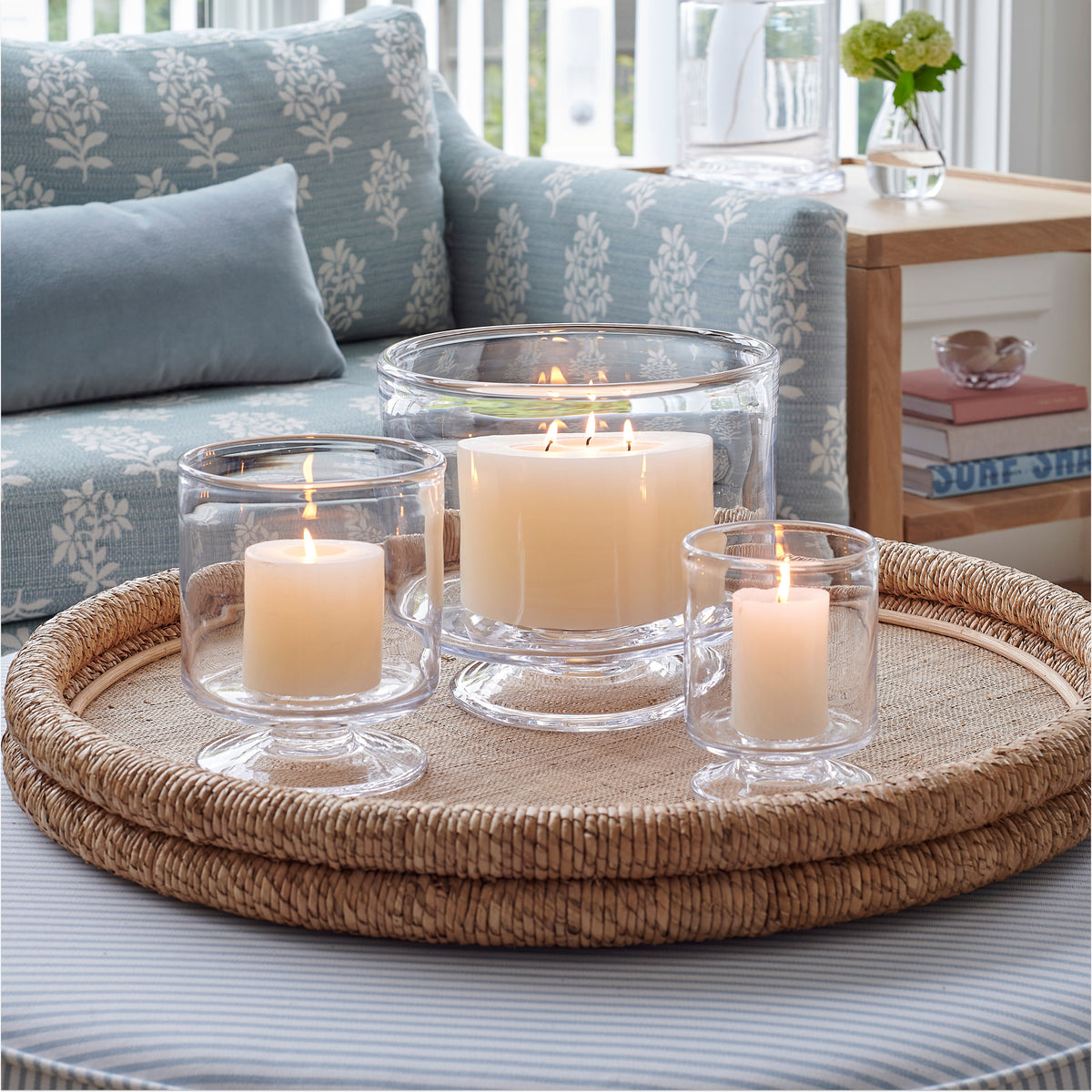 Three cream-colored pillar candles in clear glass hurricane vases arranged on a woven rattan tray.