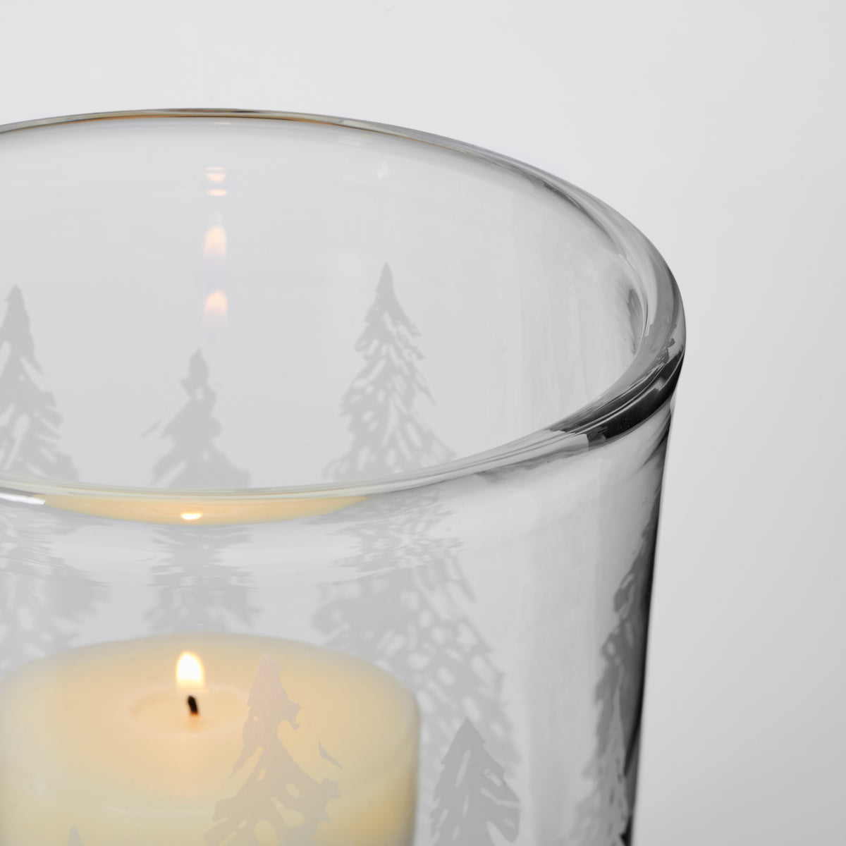 Clear glass hurricane with engraved forest tree silhouettes and cream-colored candle inside.