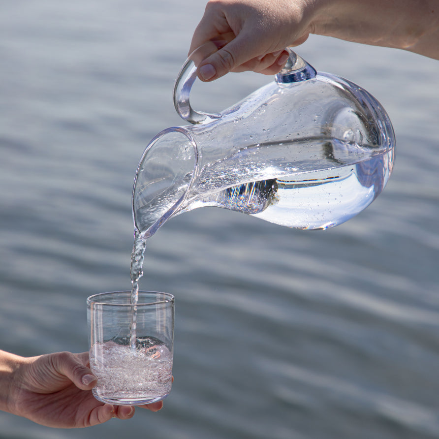 Water being poured from a Simon Pearce handblown glass pitcher into a tumbler held by a second person, with calm water in the background