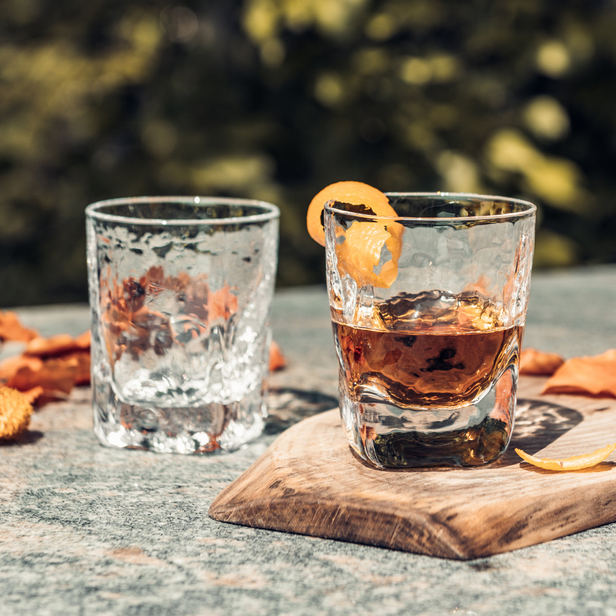 Two glasses on wooden coaster, one with clear liquid and ice, one with amber bourbon and orange peel garnish.