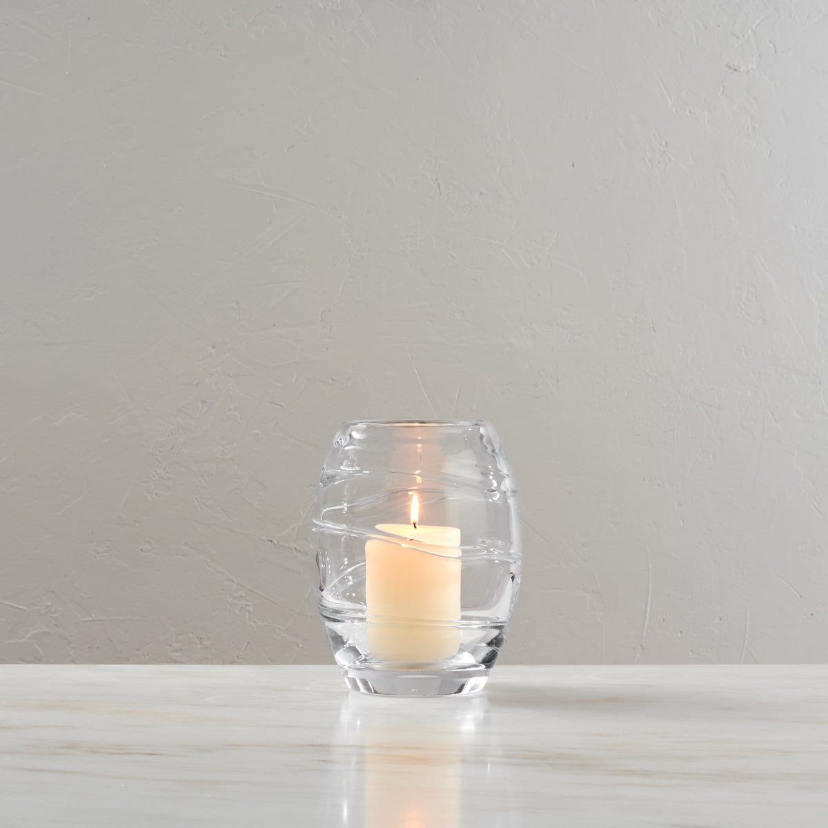 Clear glass hurricane vase with lit cream-colored pillar candle inside, displayed against a white textured background.