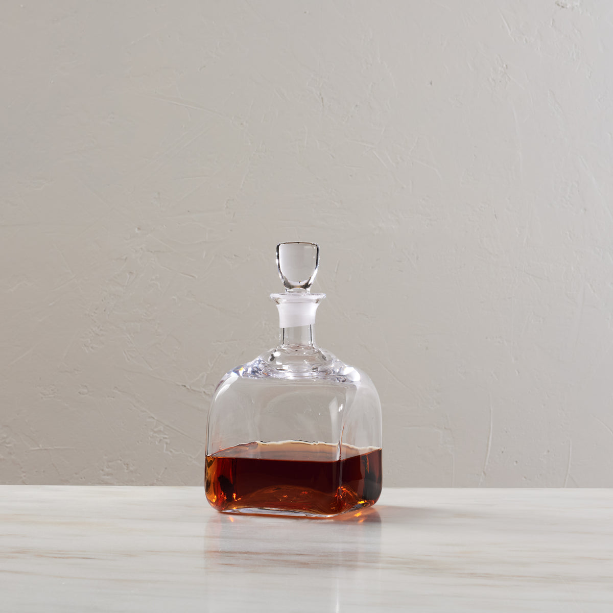 Clear glass decanter with stopper containing amber-colored liquid on white surface.