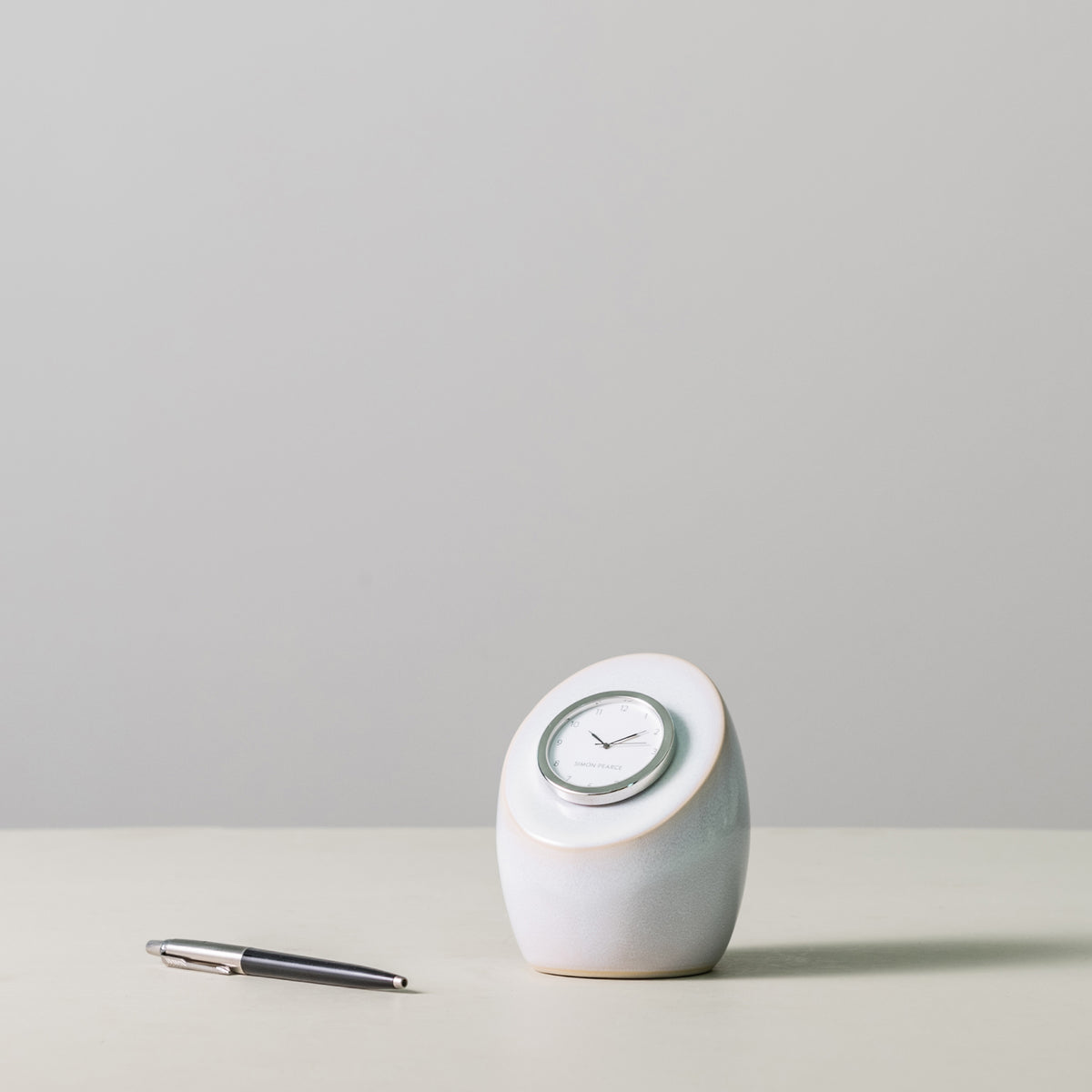 White ceramic rounded clock with gray trim and white dial face, displayed next to a pen for scale.