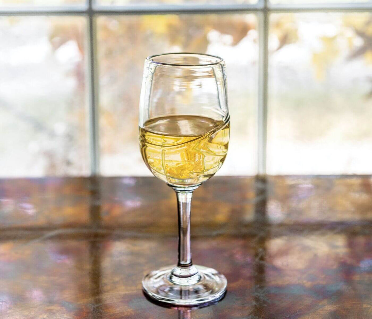 White wine in clear glass with pale yellow liquid, positioned on reflective surface near window.