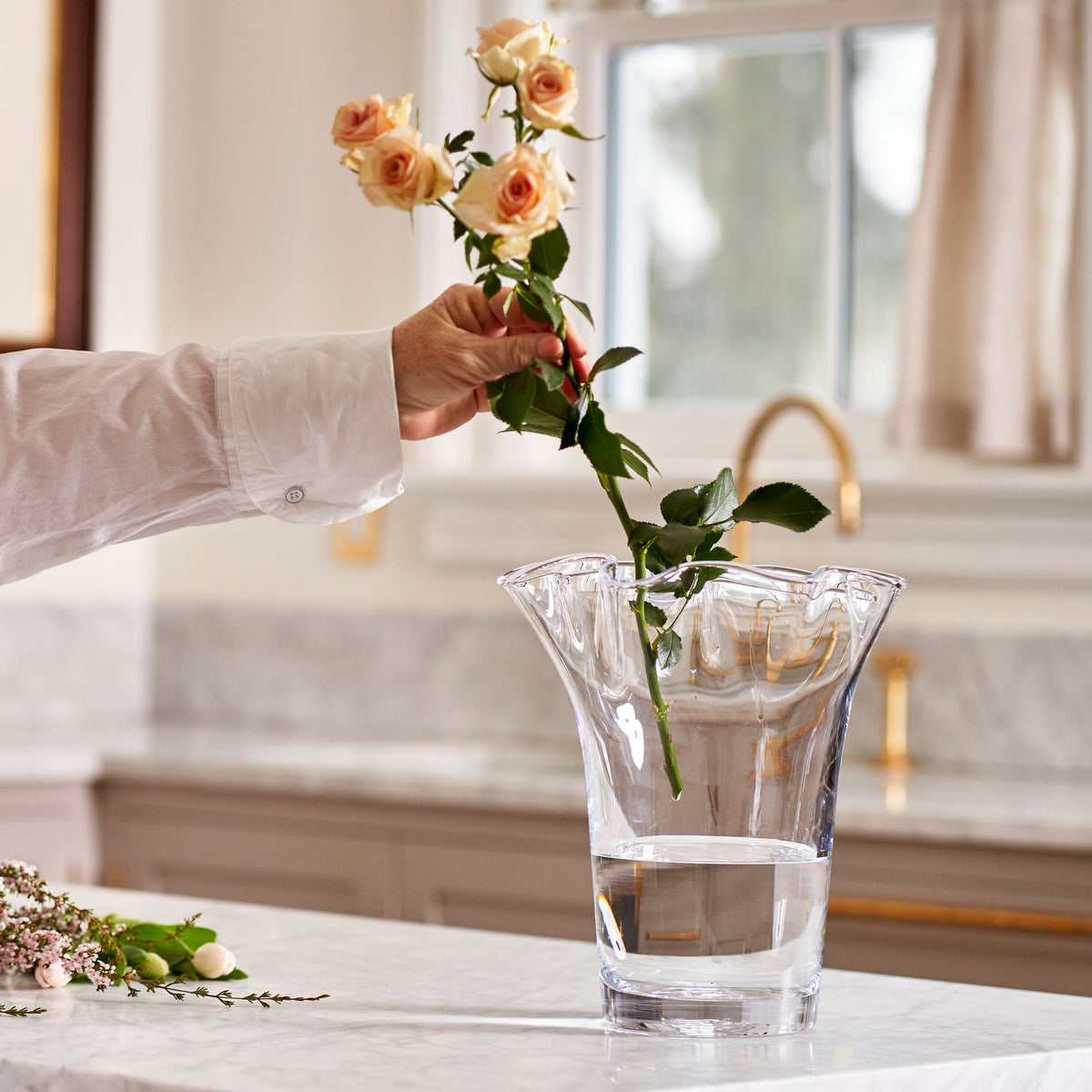 Clear glass vase with wavy edges holding peach roses and green stems, hand inserting flowers.