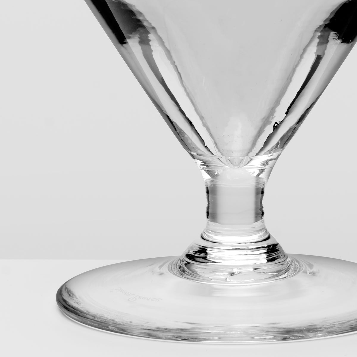 A close-up studio shot of a clear glass highball or cocktail glass base and stem on a light gray background.