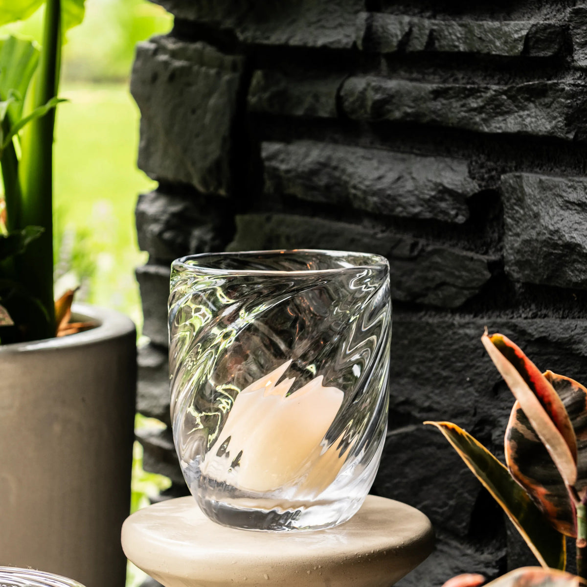 Clear glass tumbler with swirled pattern design on stone coaster, surrounded by potted plants and dark brick background.
