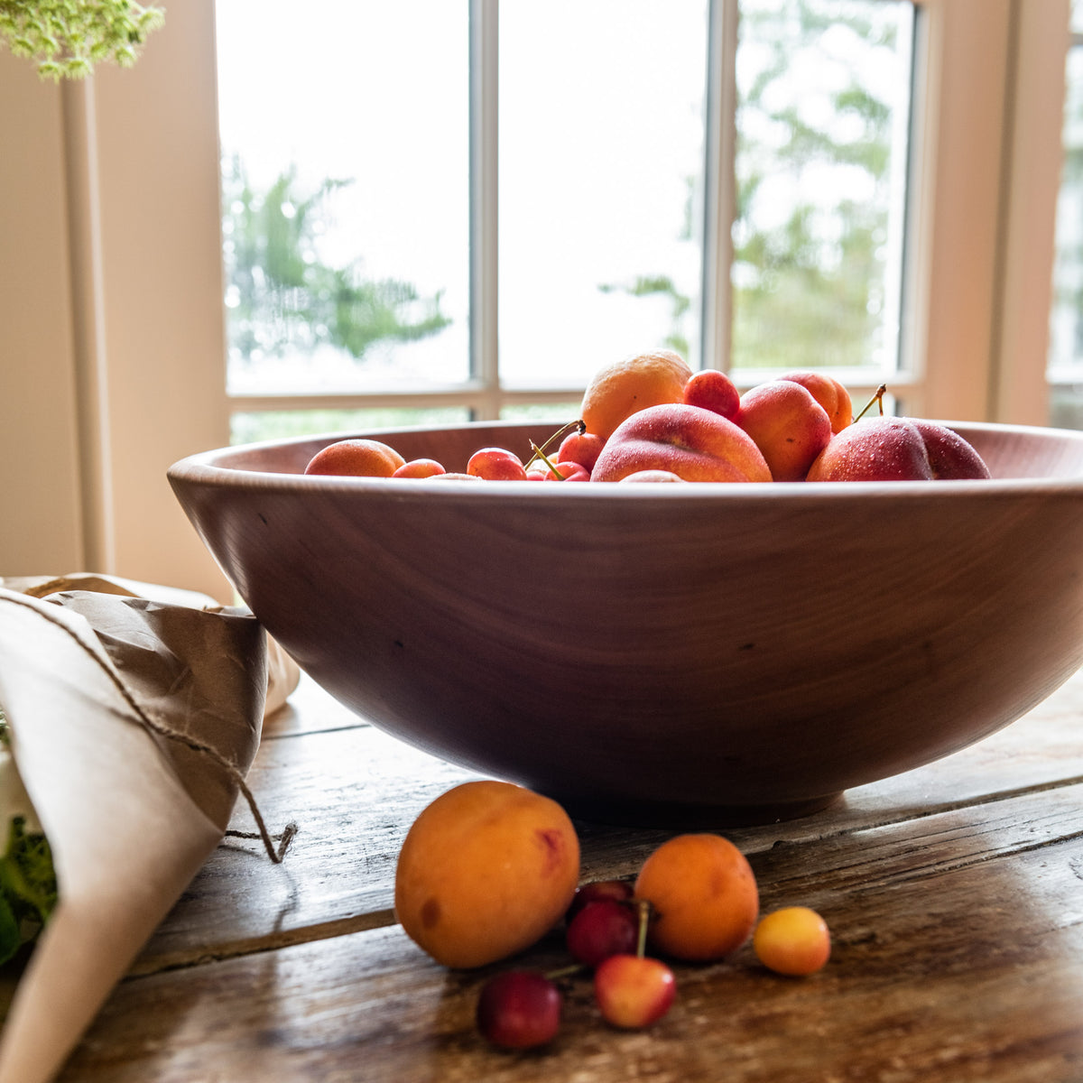 Cherry wood bowl filled with peaches and cherries on wooden table by window.