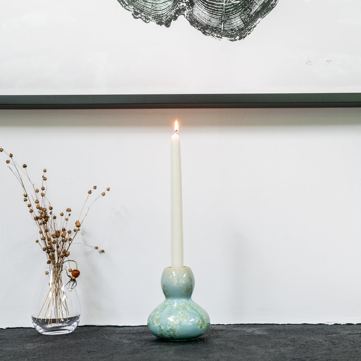 Ceramic candlestick with bulbous base in muted green glaze holding a lit cream taper candle.
