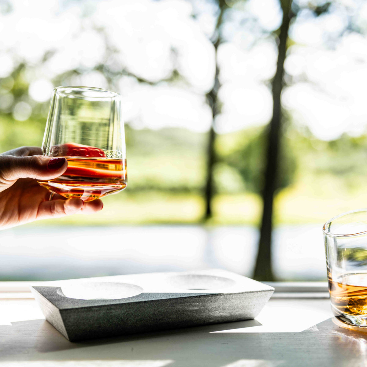 Two whiskey glasses with amber liquid, one held in hand, beside a gray soapstone base tray.