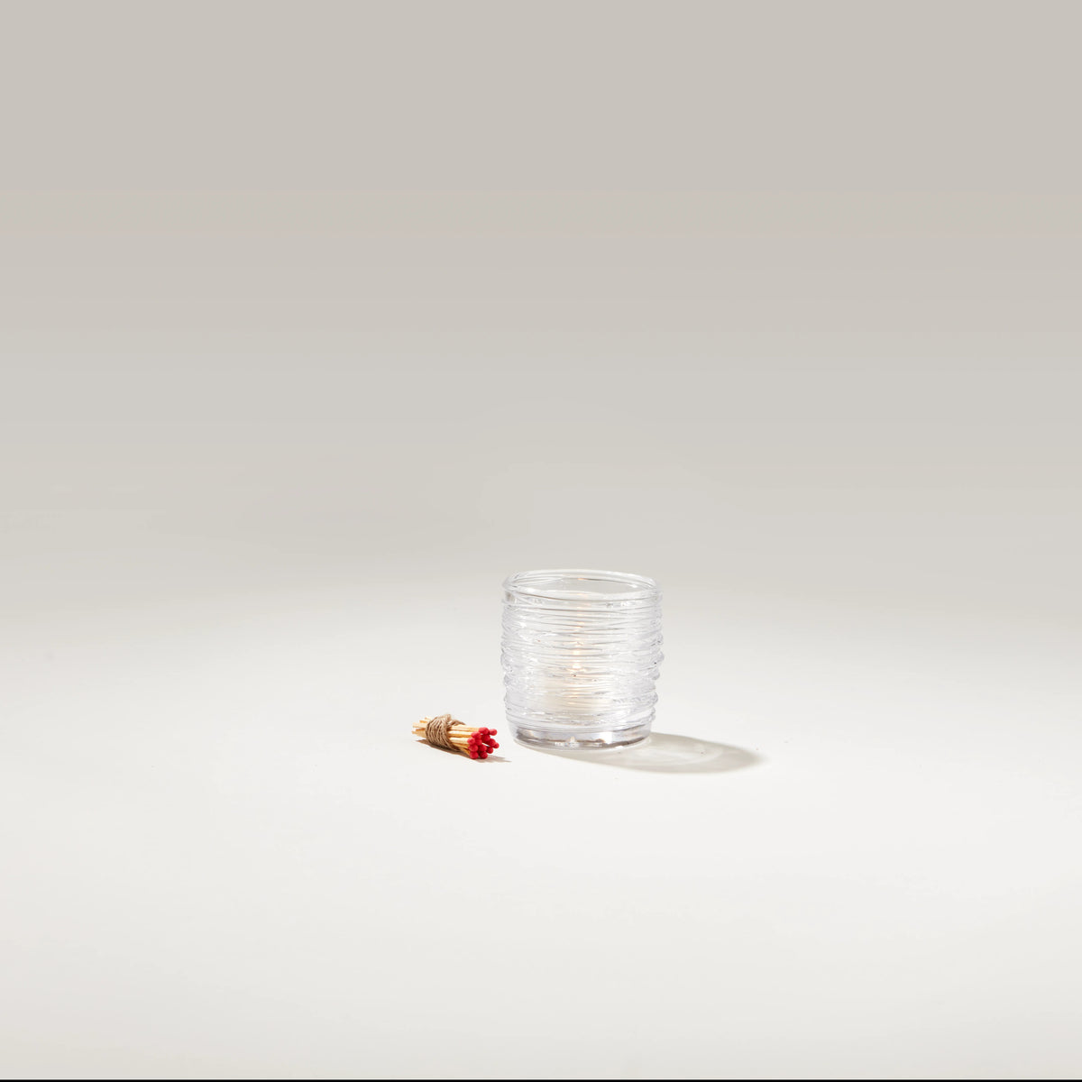 Clear glass tealight holder with textured surface and lit white candle inside, shown with red and gold match stick.