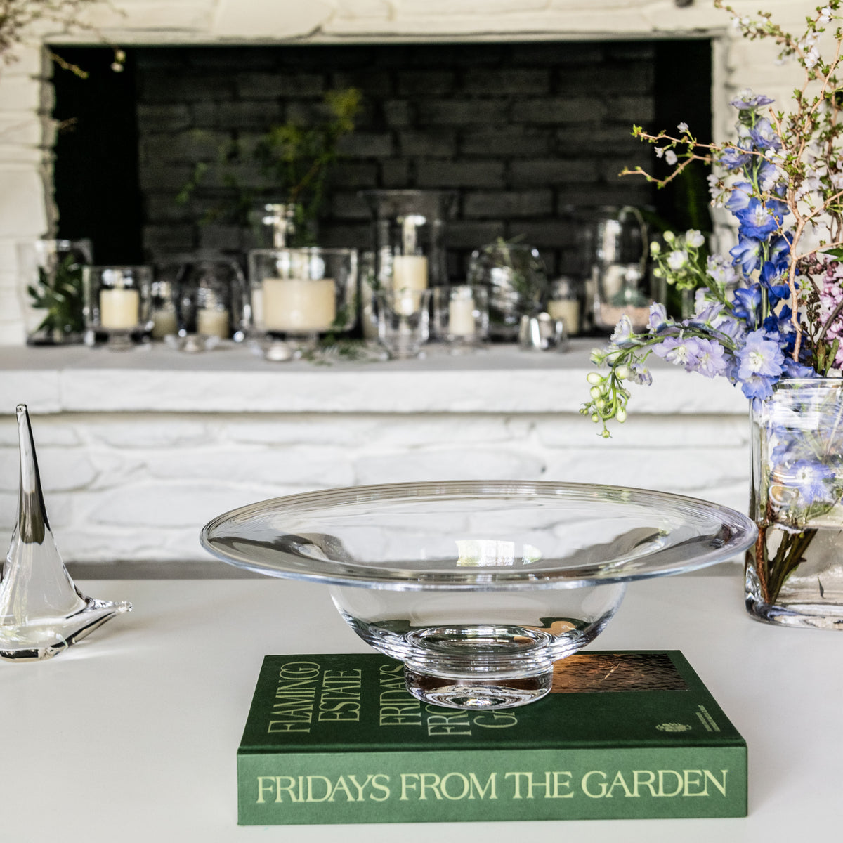Clear glass bowl with pedestal base displayed on green book, surrounded by flowers and candles.