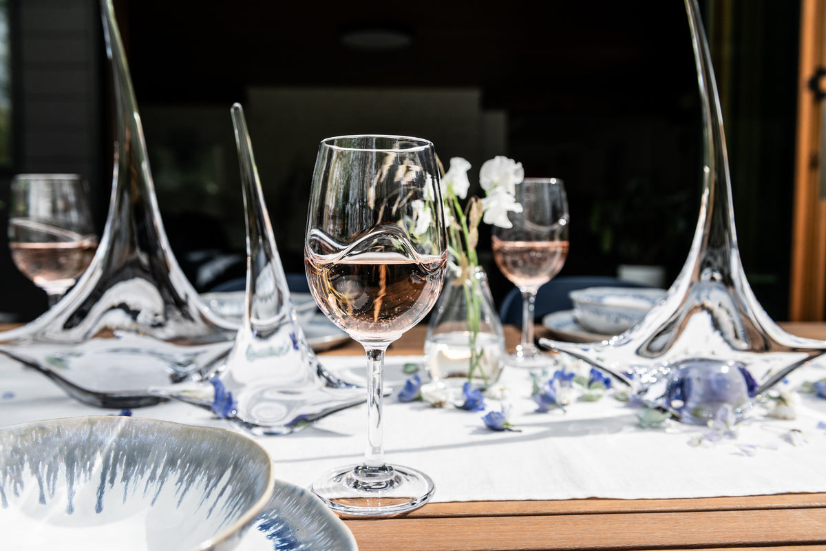 Wine glasses filled with red wine arranged on a formal dining table with place settings and white flowers.