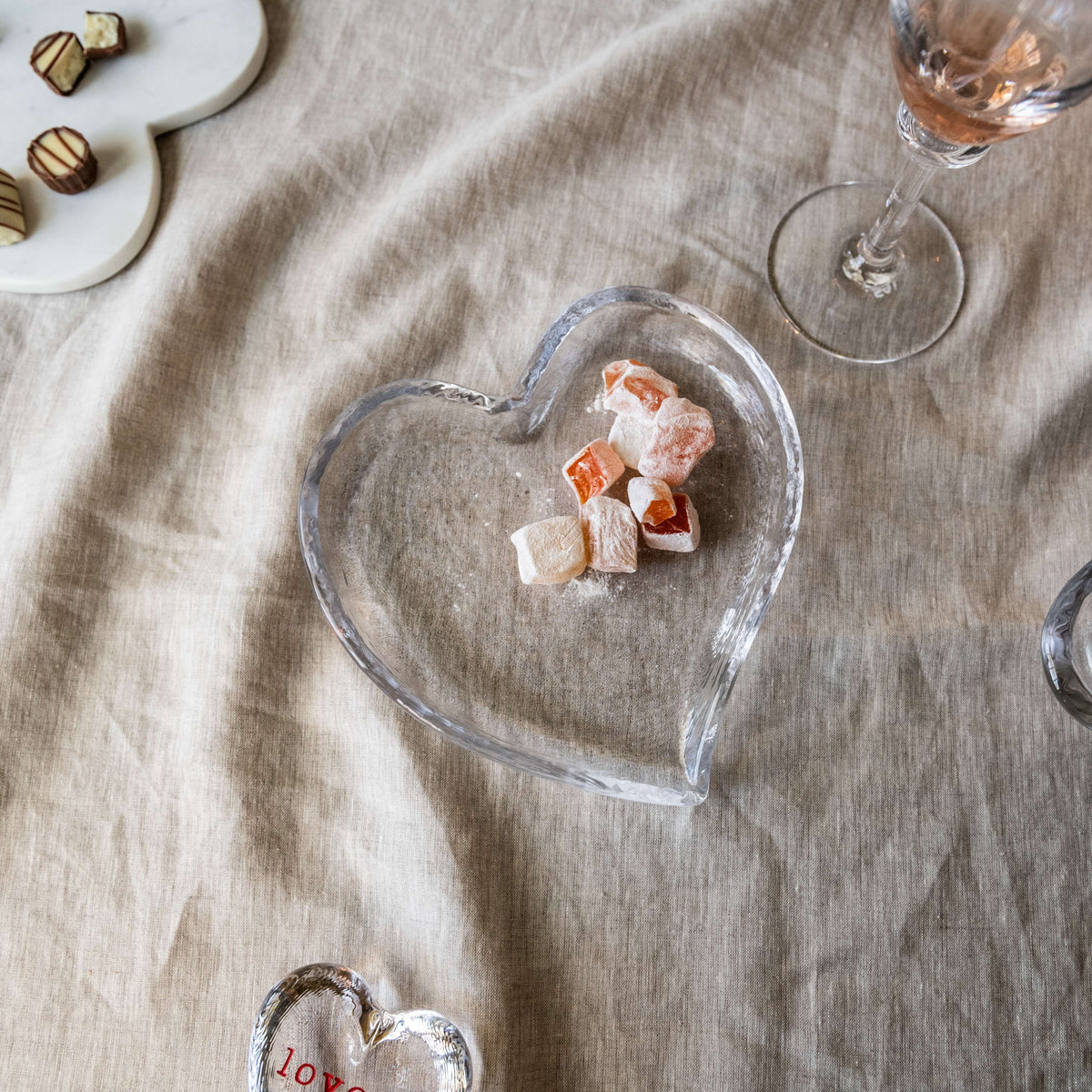 Clear glass heart-shaped tray holding pink and white candies on beige linen surface.