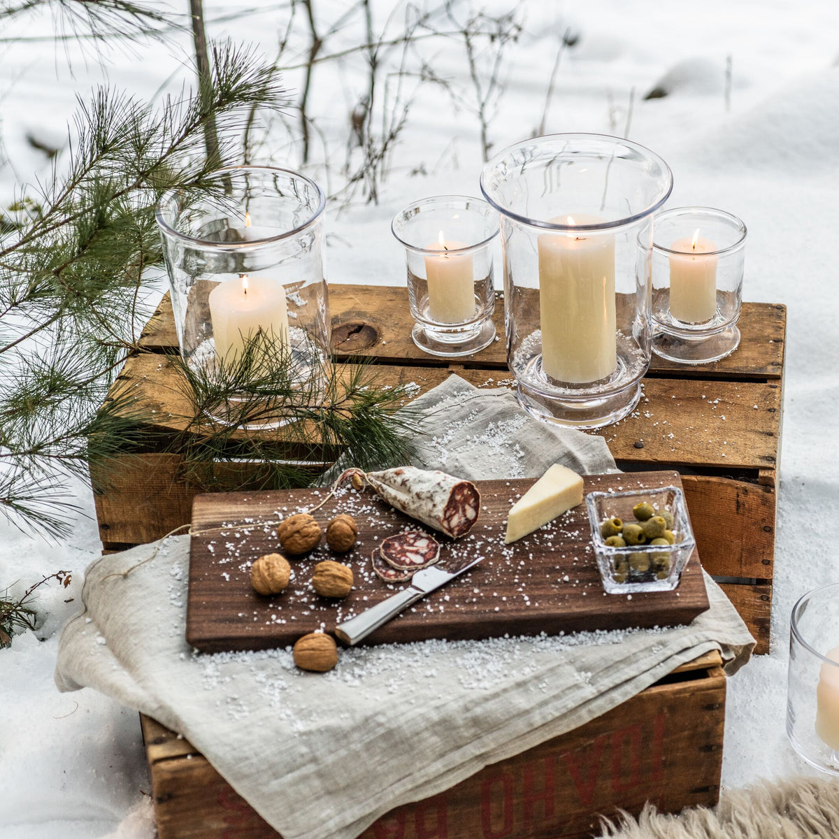 Five glass hurricane candles in varying heights arranged on wooden blocks with evergreen sprigs, cheese, cured meats, and nut