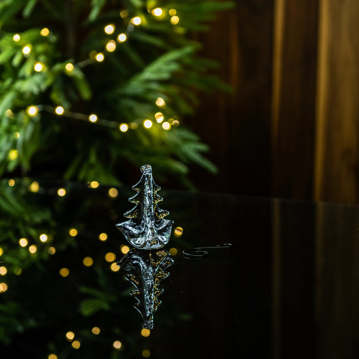 Clear glass tree-shaped ornament with gold accents, displayed in front of evergreen branches and warm white lights.