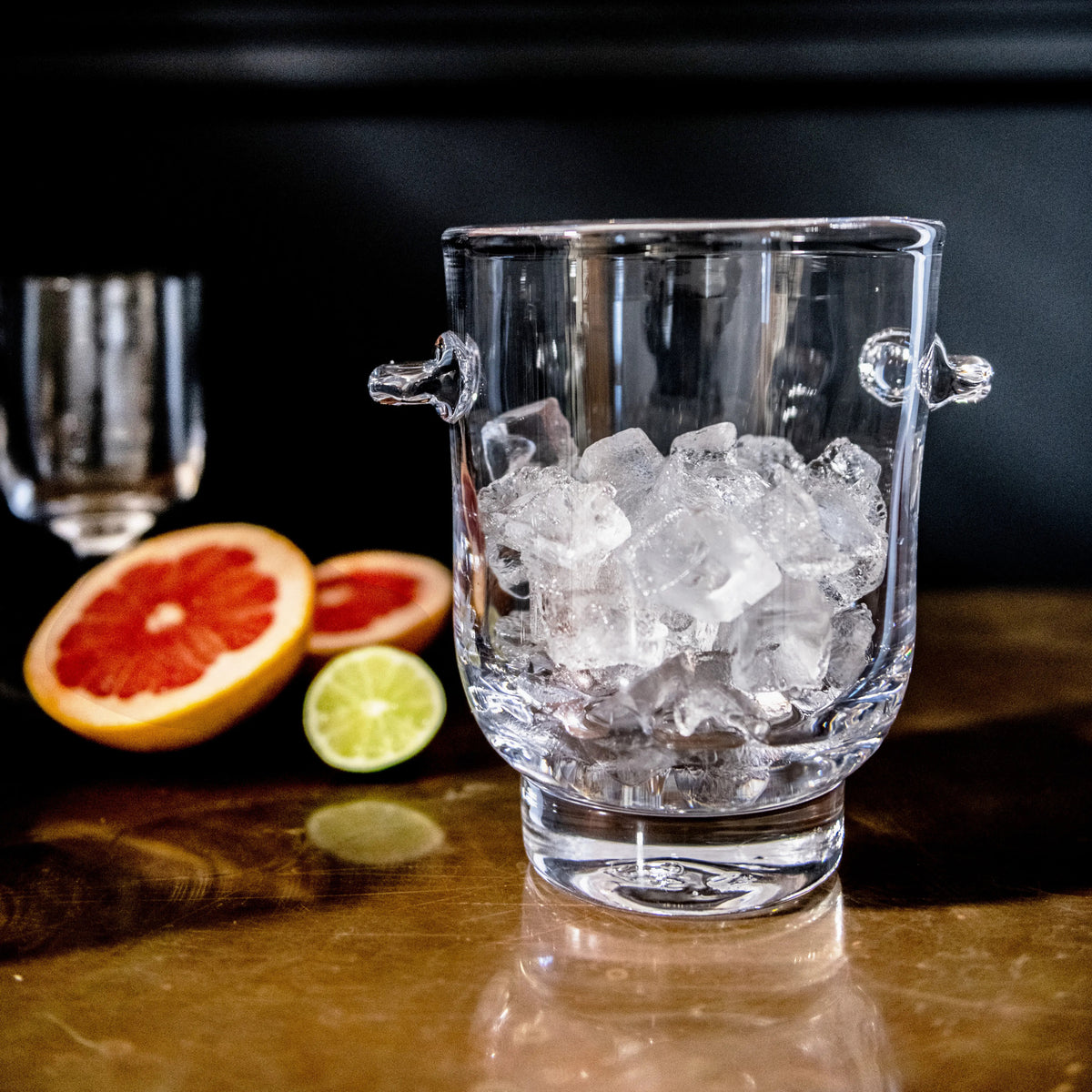 Clear glass ice bucket filled with ice cubes, dual handles, displayed with citrus slices and cocktail glass.