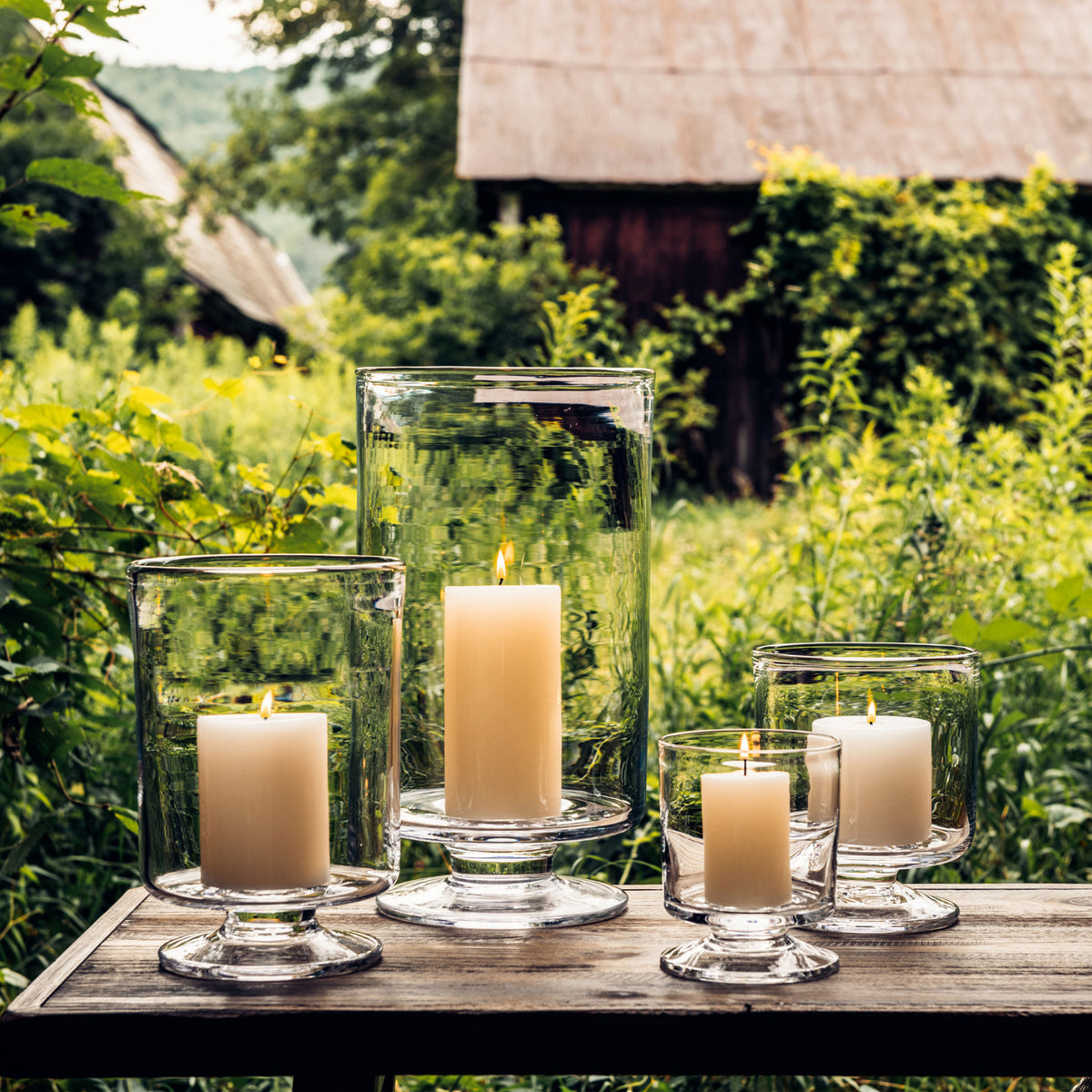 Three clear glass hurricane candleholders of varying heights with lit cream-colored candles arranged on wooden surface outdoo