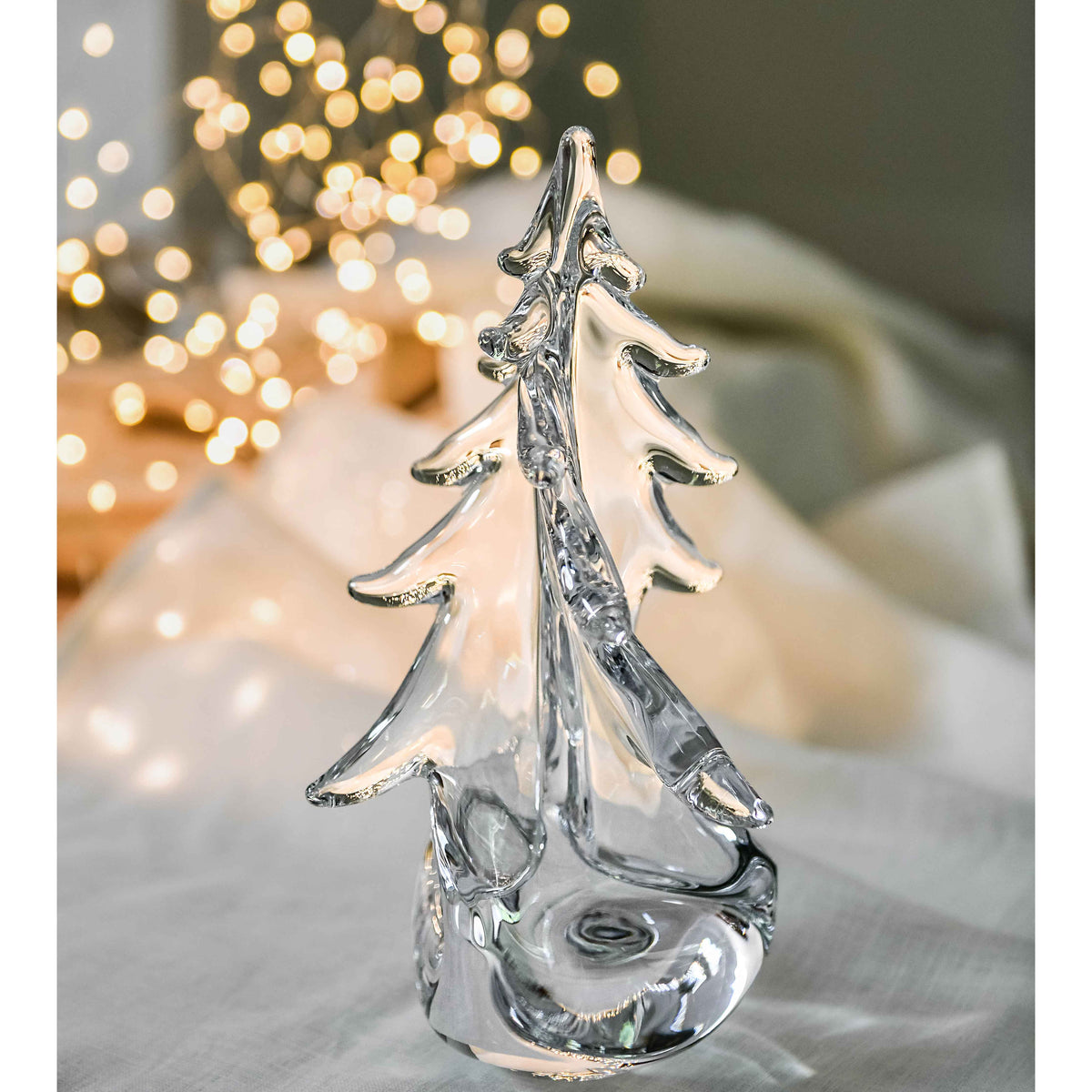 Clear glass tree-shaped figurine with twisted branches and rounded base on white surface with blurred lights behind.