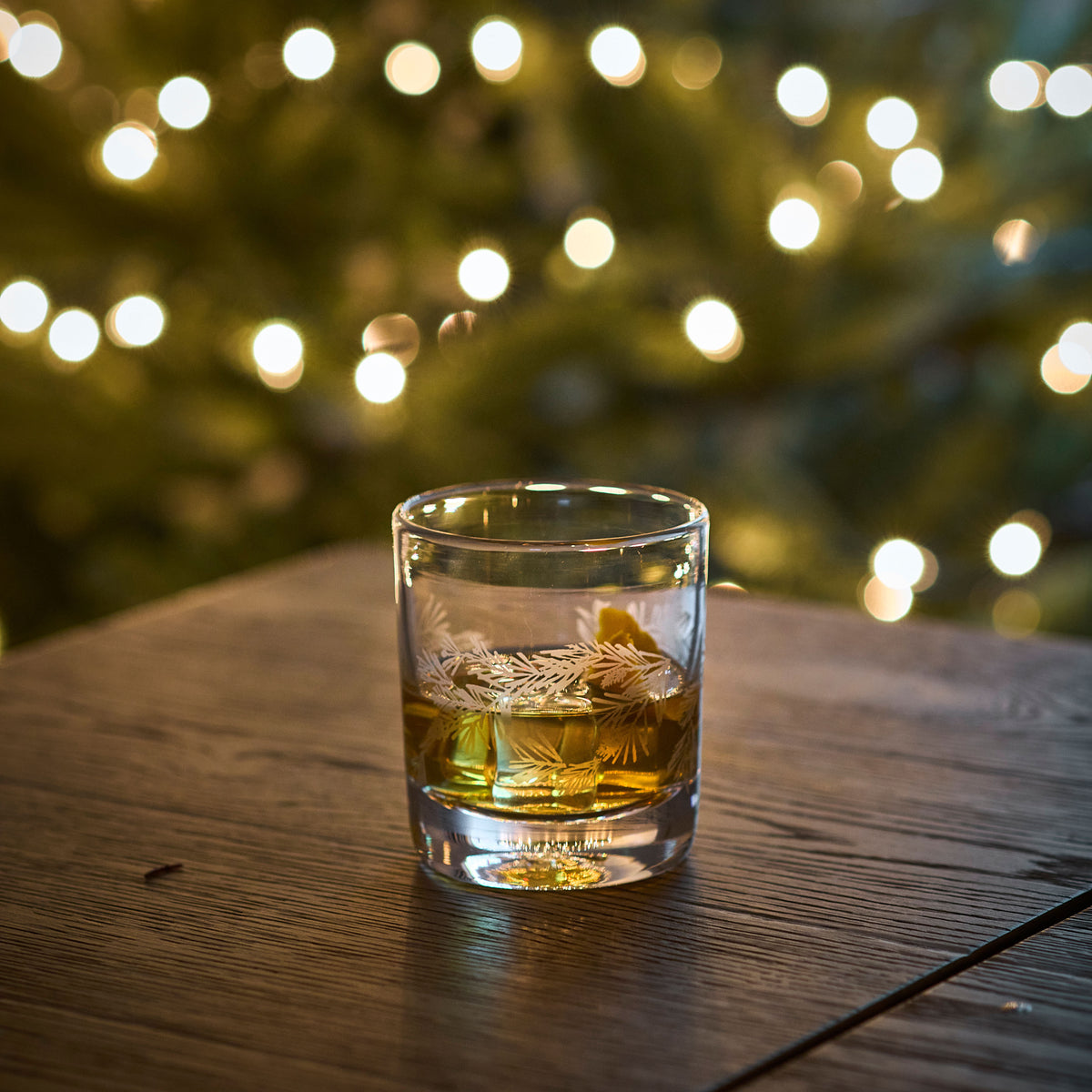 Clear double old-fashioned glass with engraved pine tree design filled with amber liquid on wooden surface.